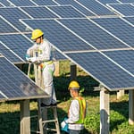 Deux techniciens en tenue de sécurité effectuent la maintenance de panneaux solaires photovoltaïques sur une installation au sol, dans un environnement verdoyant.